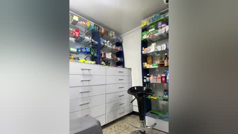 Media: Video of a small, well-lit pharmacy with white cabinets filled with colorful medicine boxes and a black stool in the foreground.