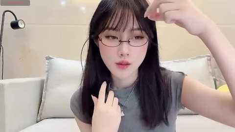 Media: Video of an East Asian woman with straight black hair, wearing glasses, adjusting her hair with a hand. She has fair skin and is dressed in a grey top. Background includes a white couch and a black desk lamp.