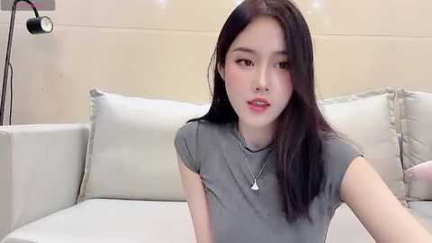 Media: Video of an East Asian woman with long black hair, fair skin, and a slender physique, wearing a grey t-shirt, sitting on a white couch.