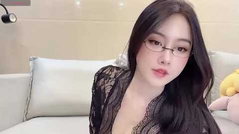 Media: Video of a young Asian woman with long black hair, wearing glasses, a black lace cardigan, and sitting on a white couch, with a stuffed toy on the right.