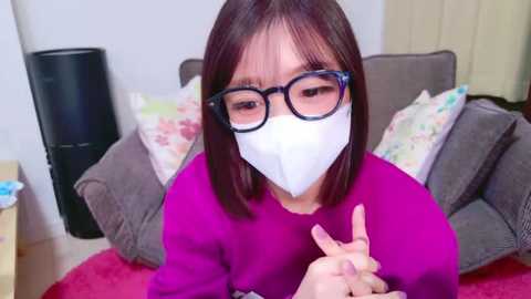 Media: Video of an Asian woman with straight brown hair and glasses, wearing a white mask, purple shirt, and beige sofa.