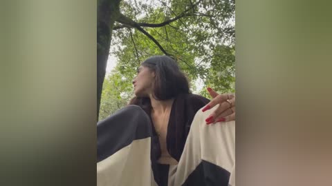 Media: A video of a young woman with long brown hair, wearing a dark robe, sitting in a natural, green-lit forest setting. Her red-painted nails are visible, and she gazes thoughtfully at the camera.