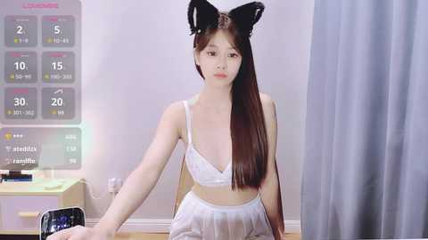 Media: Video of an East Asian woman with long brown hair, wearing a white lingerie set and black cat ears, sitting on a wooden floor in a minimalist room with a digital clock and grey curtains.