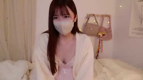 Media: Video of a young Asian woman with long brown hair, wearing a white mask and a loose white robe, sitting on a bed with white sheets in a cozy, dimly lit room.