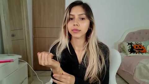 Media: Video of a young Asian woman with long, straight blonde hair and fair skin, wearing a black top, playing with her hands in a cozy bedroom with light wood furniture, a bed, and a pumpkin pillow.