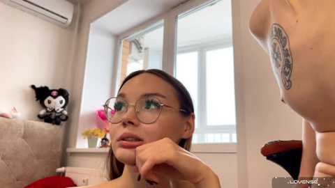 Media: Video of a young woman with glasses, long brown hair, and a small tattoo on her arm, sitting on a couch, looking at a large stuffed animal. Background includes a window and plush toys.