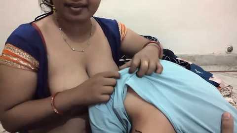 Media: Video of a South Asian woman with medium brown skin, wearing a blue saree with gold and orange borders, exposing her large breasts, holding a light blue cloth, in a simple indoor setting.