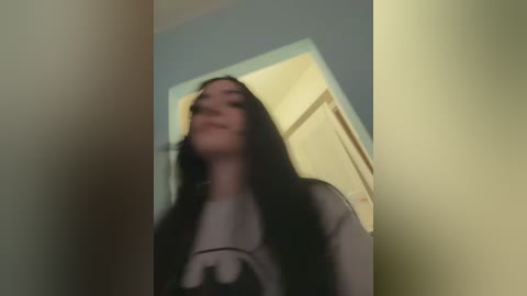 Media: A blurry video of a young woman with long black hair, wearing a grey t-shirt, standing in a dimly lit room with a yellow door and blue wall in the background.