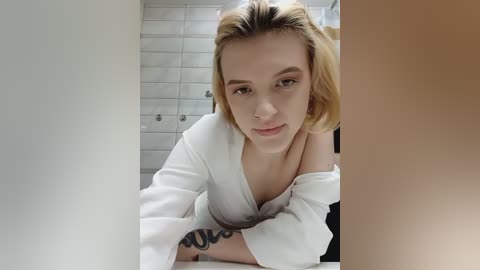 Media: Video of a young, light-skinned woman with short, blonde hair, wearing a white off-shoulder top. She has a tattoo on her right forearm and is leaning over a table. Background shows white tiled walls.