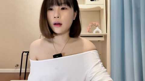 Media: Video of an Asian woman with straight, shoulder-length brown hair and a white off-shoulder top, seated indoors. She wears a pendant necklace. Background includes a light beige wall, wooden table, and a pink vase.