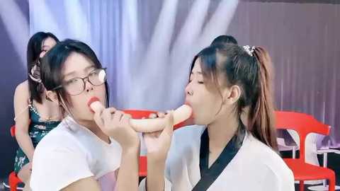 Media: Video of two young Asian women in glasses, wearing white tops, sucking on a large, phallic object, set against a studio backdrop with a red chair and grey curtains.