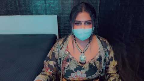 Media: Video of a South Asian woman with a light brown complexion, wearing a floral saree, gold jewelry, and a blue face mask, sitting on a dark bed against a textured wall.