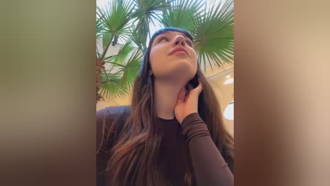 Media: Video of a young woman with long brown hair, wearing a black top, gazing upwards with a contemplative expression, surrounded by lush green palm fronds.