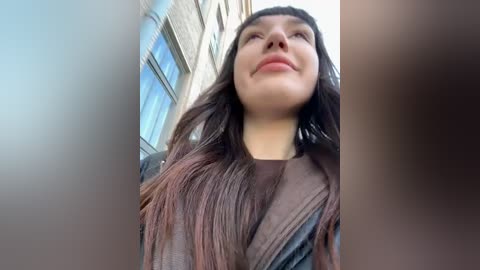 Media: A video of a young woman with long, dark hair and a slight smile, wearing a brown, textured top, standing outdoors near a building with large windows.