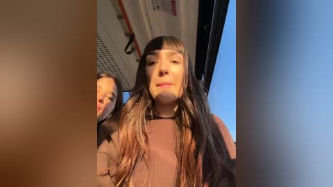 Media: Video of two young women with long hair, one in a brown sweater, the other in a black sweater, taken from the front inside a bus, with sunlight streaming in, creating a warm, intimate atmosphere.