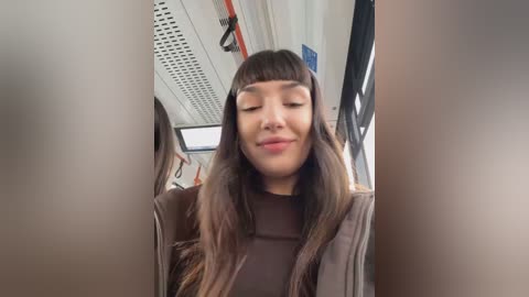 Media: Video of a smiling Asian woman with long, straight brown hair, wearing a brown sweater, seated in a subway car with beige walls and blue signs.