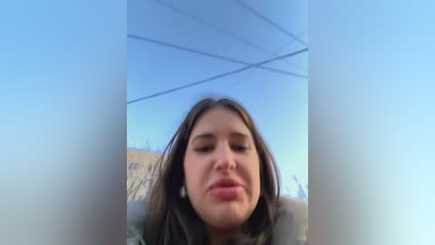 Media: Video of a young woman with long brown hair and closed eyes, standing outdoors in daylight with a clear blue sky, wearing a green jacket.