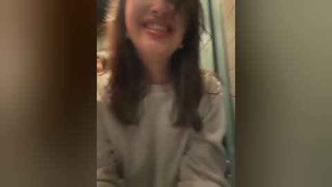 Media: Video of a young woman with light brown hair and a wide smile, wearing a white shirt, in a dimly lit bathroom with a green shower curtain.