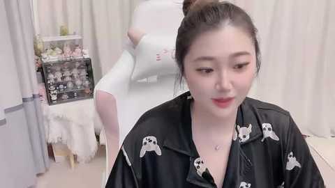 Media: Video of an Asian woman with light skin and dark hair in a bun, wearing a black robe with white panda prints, seated in a softly lit bridal salon with white curtains and floral arrangements.