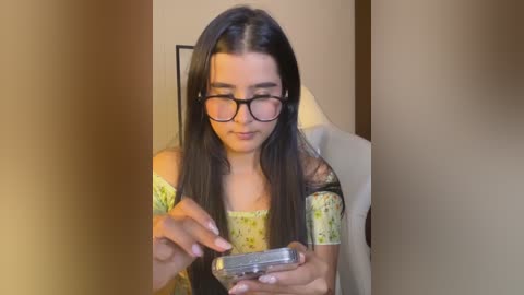 Media: Video of a young woman with long dark hair, wearing black-rimmed glasses, a yellow floral off-shoulder top, and a gray blazer, holding a silver phone, indoors with a beige wall.