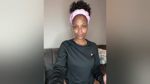 Media: Video of a Black woman with a medium-dark skin tone, wearing a pink headband, black long-sleeve shirt, and seated on a dark couch.