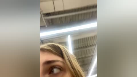 Media: Video of a blonde woman with light skin, her face partially visible, looking upward. Background features an industrial ceiling with metal beams and fluorescent lights.