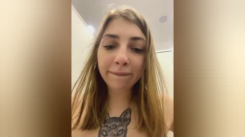 Media: Video of a young Caucasian woman with long, straight blonde hair, wearing a minimalist outfit, showcasing a large, detailed tattoo of a cat's face on her chest. Background is a neutral indoor setting.