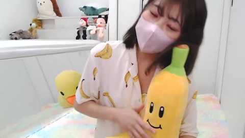 Media: Video of an Asian woman with straight black hair, wearing a white mask, and a white shirt with cartoon fruit patterns, hugging two plush bananas. Background features a white bathtub and plush toys, creating a playful, whimsical atmosphere.