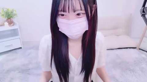 Media: A video of an Asian woman with long, straight black hair, wearing a white mask, white blouse, and a white skirt, kneeling on a soft, light-colored carpet in a minimalist, white-walled room with a white chair and a white nightstand with a small plant.