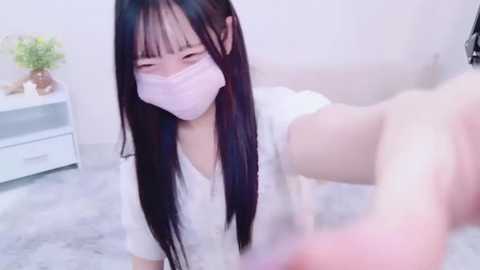 Media: Video of an Asian woman with long black hair, wearing a white mask and white shirt, kneeling on a fluffy carpet in a minimalist, light-colored room.