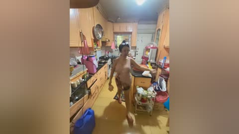 Media: Video of a half-naked woman with small breasts, in a messy kitchen, holding a pink item, surrounded by cluttered wooden cabinets and appliances.