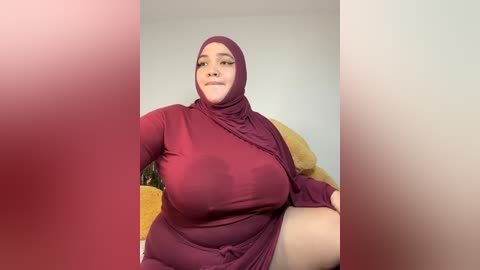 Media: A video of a plus-sized woman with fair skin, wearing a maroon hijab and dress, sitting on a bed with plush toys, smiling confidently.