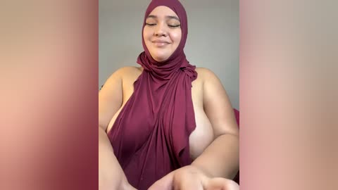 Media: Video of a plus-sized woman with medium brown skin, wearing a maroon hijab and a matching maroon top, revealing her large, natural breasts. She has a content expression and is seated against a plain, light-colored background.