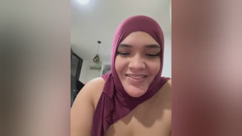 Media: Video of a smiling woman with medium brown skin, wearing a maroon hijab and a draped maroon top, standing indoors with blurred background.