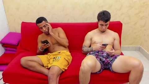 Media: Video of two shirtless men, one in yellow shorts, the other in patterned boxers, sitting on a red couch, engrossed in smartphones, against a beige wall.