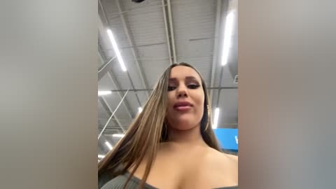 Media: Video of a smiling, light-skinned woman with long, straight brown hair, wearing a grey off-shoulder top, in a warehouse with exposed pipes and fluorescent lights.