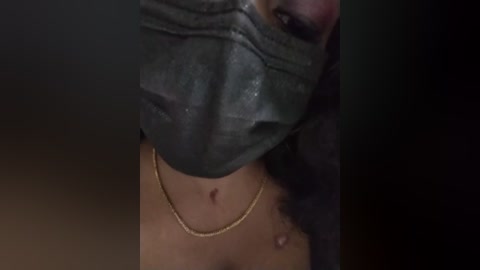 Media: A close-up video of a woman wearing a grey surgical mask and a gold necklace, with visible moles on her neck, in a dimly lit room.