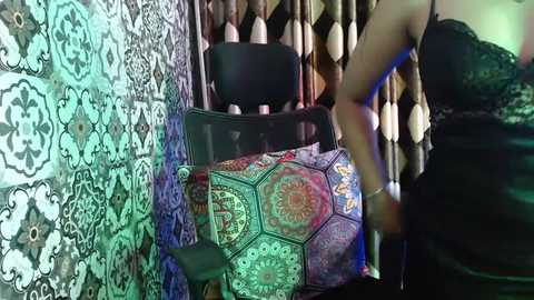 Media: Video of a woman in a black lace top standing beside a black chair covered with a vibrant, intricate mandala-patterned blanket in a room with patterned, colorful wallpaper and hanging beaded curtains.