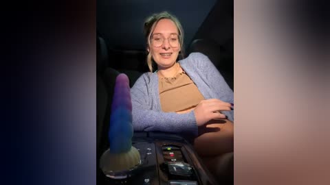 Media: Video of a blonde woman with glasses, light skin, and a beige top, sitting in a car, holding a multi-colored dildo and smiling.