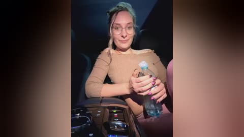 Media: Video of a woman with fair skin and glasses, wearing a beige top, holding a water bottle in a dimly lit car, with a black steering wheel in the foreground.