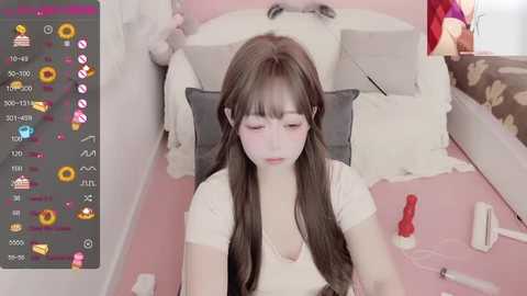 Media: A video of a young Asian woman with long brown hair and fair skin, sitting at a desk with a pink background, surrounded by various items including a phone, a red bottle, and a black pillow.