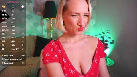 Media: A video of a blonde woman with short hair, wearing a low-cut red dress, smiling and winking in a dimly lit room with green lighting and a black lamp.