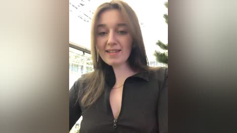 Media: Video of a young woman with straight, shoulder-length light brown hair, wearing a black, unbuttoned shirt. She stands in an indoor setting with a blurred background of a building and a tree.