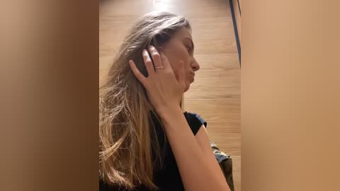 Media: Video of a young woman with light skin and long, wavy blonde hair, wearing a black top, standing in a wooden hallway, holding a phone to her ear.