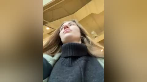 Media: A video of a young woman with straight brown hair, wearing a black turtleneck sweater, standing in a dimly lit, beige-hued room with a wooden ceiling and large windows.