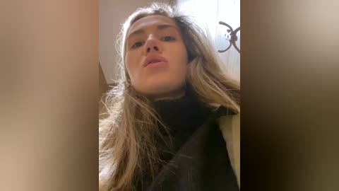 Media: Video of a young Caucasian woman with light brown hair and pale skin, wearing a black turtleneck sweater. She has a neutral expression and is standing in a dimly lit room with a blurred background.