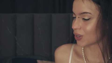 Media: Video of a young woman with fair skin, dark hair, and a slender physique, wearing a pearl necklace. She is in a dimly lit room with dark curtains, looking contemplative.