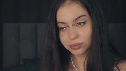 Media: Video of a young woman with long, straight black hair, fair skin, and subtle makeup. She wears a simple pearl necklace. The background is a dimly lit room with a tufted headboard.