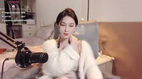 Media: A video of an Asian woman with pale skin, long black hair, and medium-sized breasts, sitting in a cozy room with a white furry coat, recording herself on a camera.