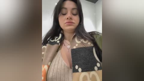 Media: Video of a young woman with long, dark hair, wearing a beige knit sweater with a black and white pattern, looking serene in a softly lit room.